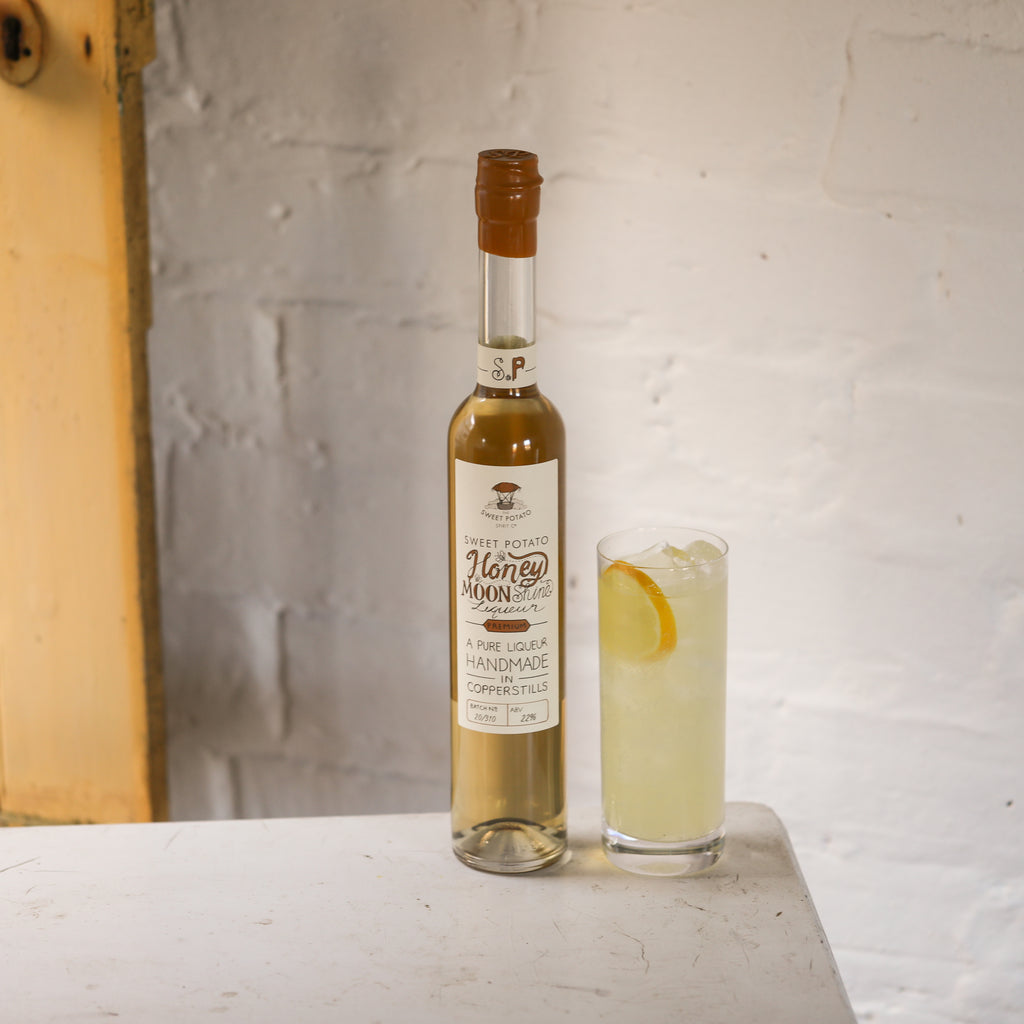 bottle of honey moonshine with a lemonade drink next to it in a neutral white brick background. sweet potato spirit, sweet potato, spirit, sweet, honey, moonshine, honey moonshine, liqueur, honey moonshine liqueur, desert, gifting, spirits